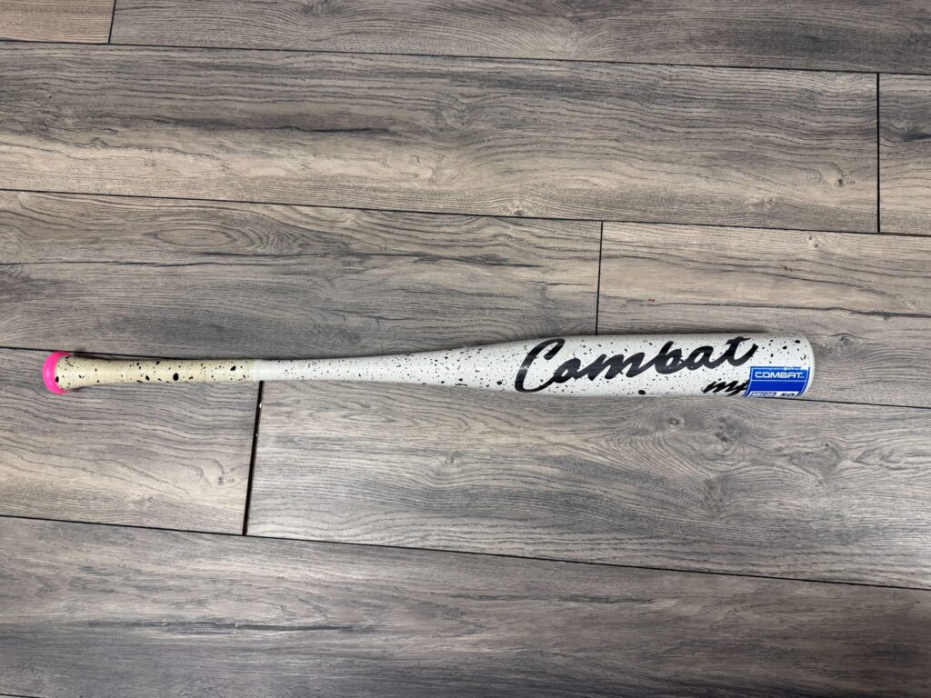 A limited edition white and black speckled Combat BBCOR baseball bat called Cookies and Cream resting on a wooden floor, representing high school travel ball equipment expenses.