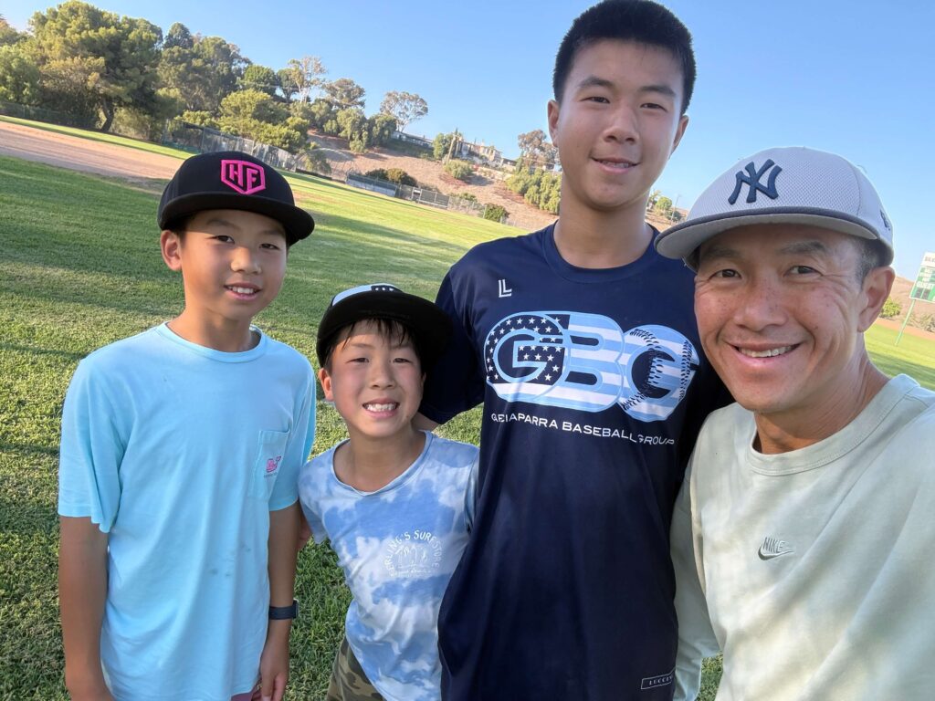 Travel baseball dad Simon Chan taking a selfie with his three sons at a local baseball field, representing a family navigating the costs of youth travel baseball.