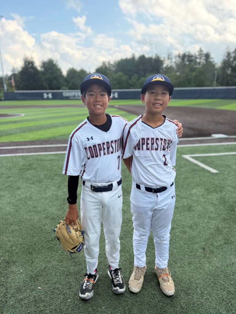 Brendan and Christian stand with their arms around each other on a turf baseball field, both smiling in their white Cooperstown All Star Village uniforms.