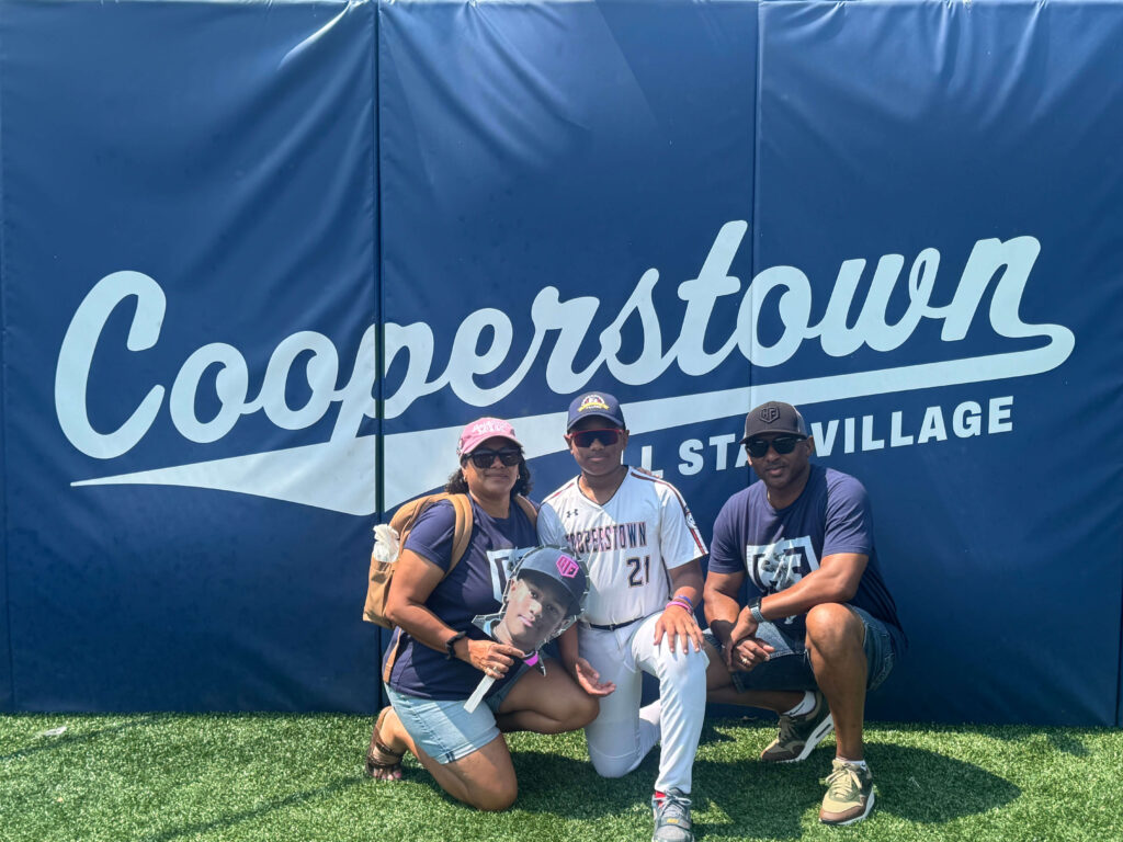 The Chris Henry family poses together in front of a blue Cooperstown All Star Village wall, with their son kneeling in his baseball uniform between his parents.