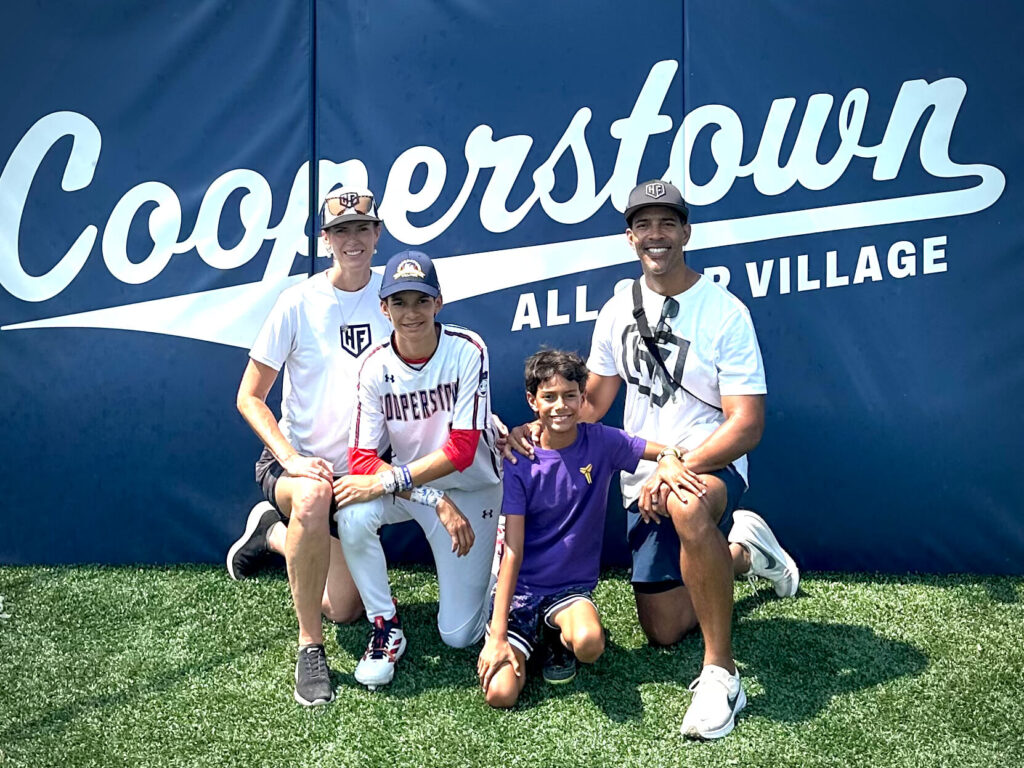 The Kennon Jackson family poses together in front of a blue Cooperstown All Star Village wall, with their son kneeling in his baseball uniform alongside his parents and brother.