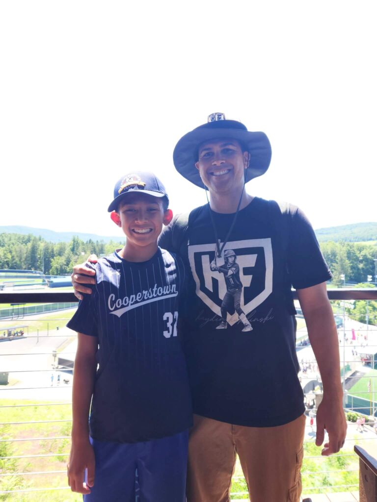 Mike Kuklinski stands with his arm around a smiling young player in a blue Cooperstown jersey on a balcony overlooking the Cooperstown All Star Village baseball fields.