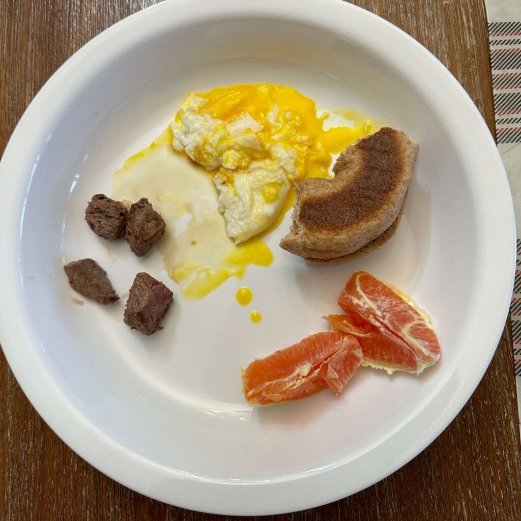 White plate with eggs, half a bagel, some steak chunks and grapefruit