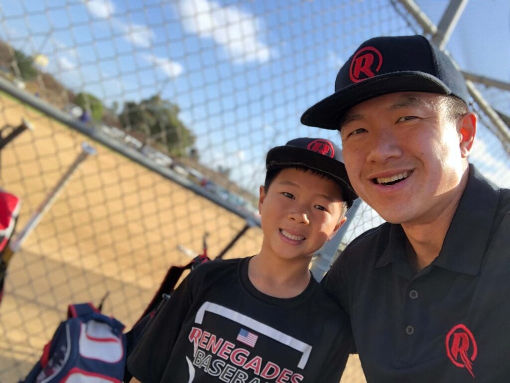 A travel baseball coach and his son taking a smiling selfie by the dugout fence, wearing matching black and red Renegades team hats.