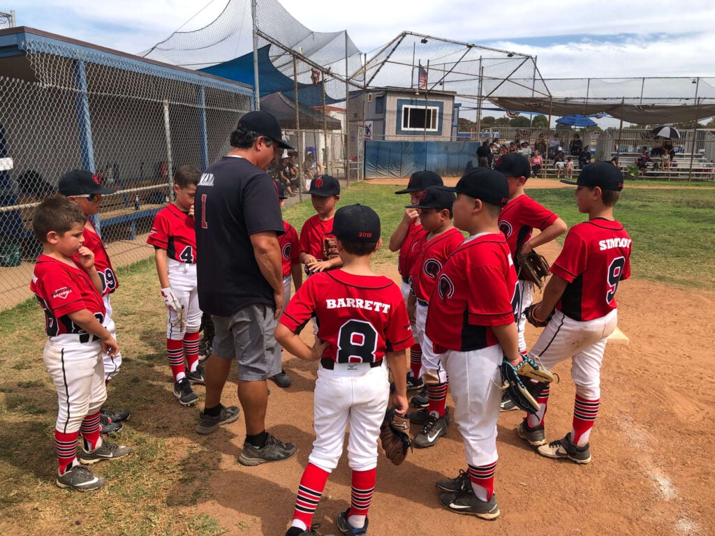 A youth travel ball team in red uniforms gathered on the dirt infield, listening attentively to their coach after practice.