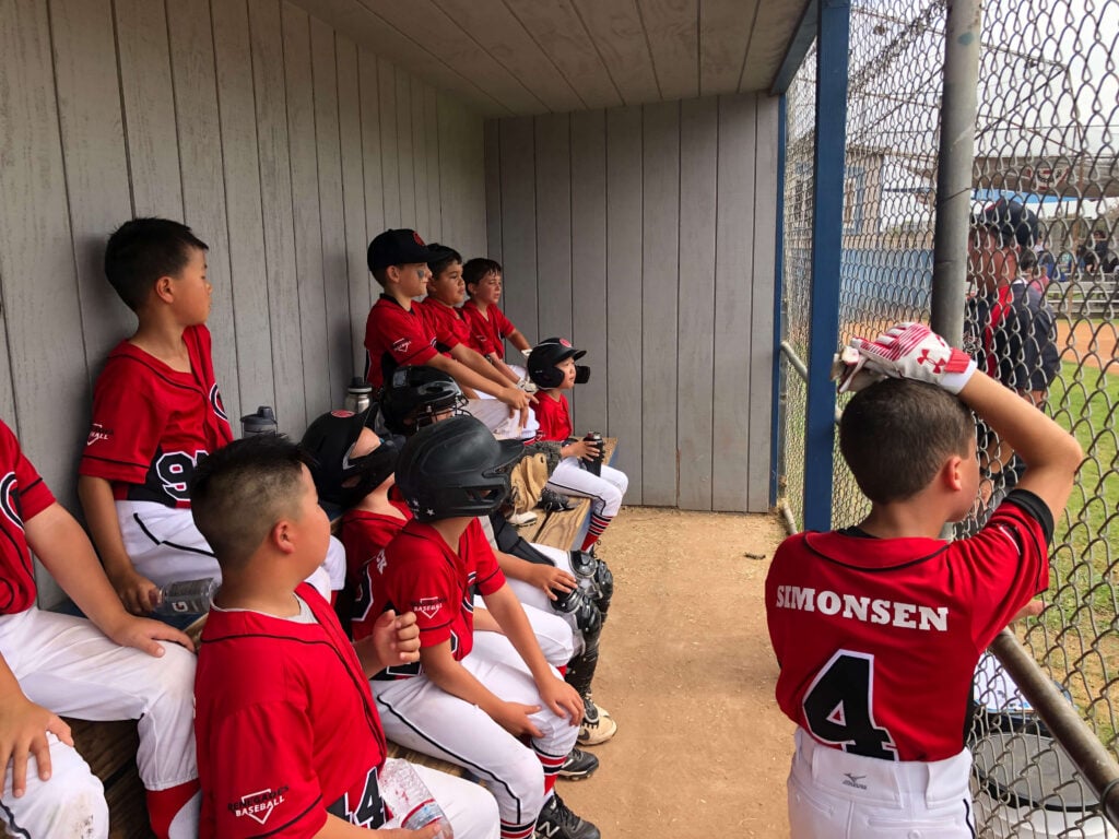 Young travel ball players sitting on a wooden bench in the dugout, watching the game action through the chain-link fence.