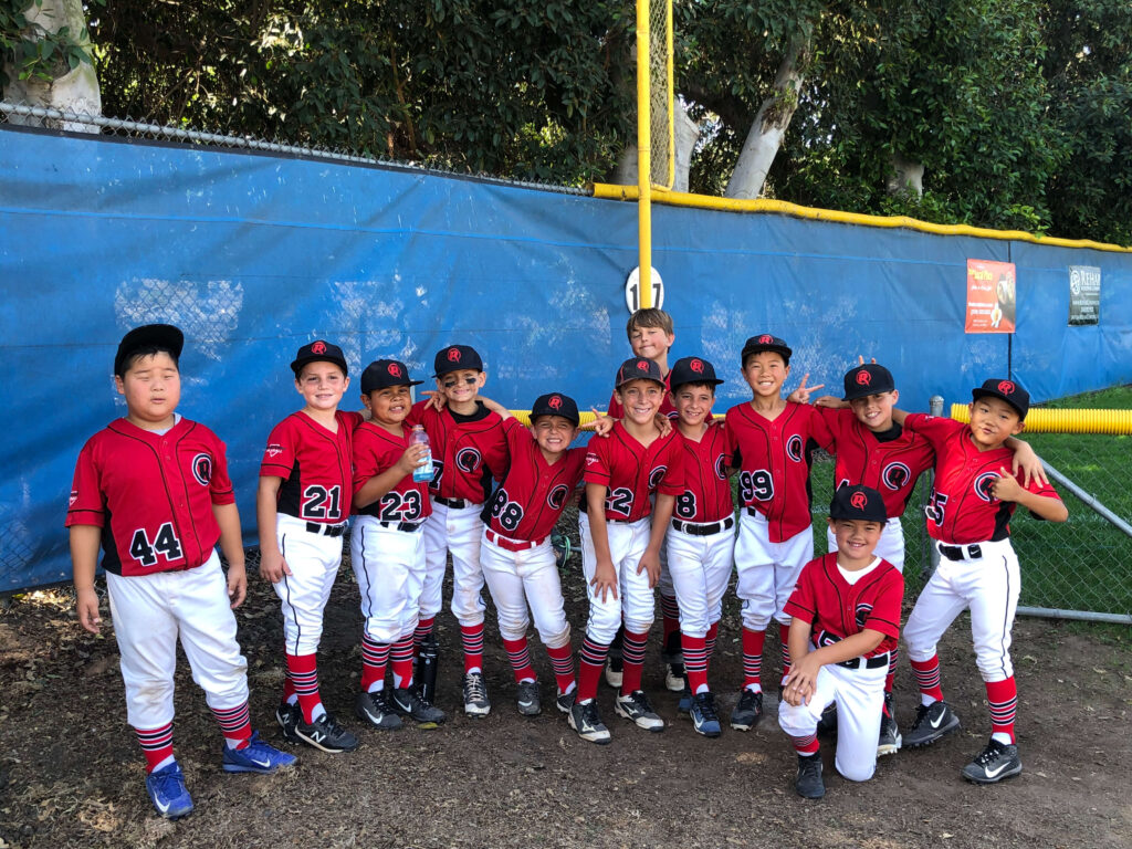 The Renegades youth travel baseball team smiling for a group photo in their red and black uniforms in front of a blue outfield windscreen.