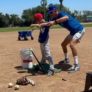 A travel baseball coach providing 1-on-1 pitching instruction to a young player on an outdoor dirt field, demonstrating focused player development