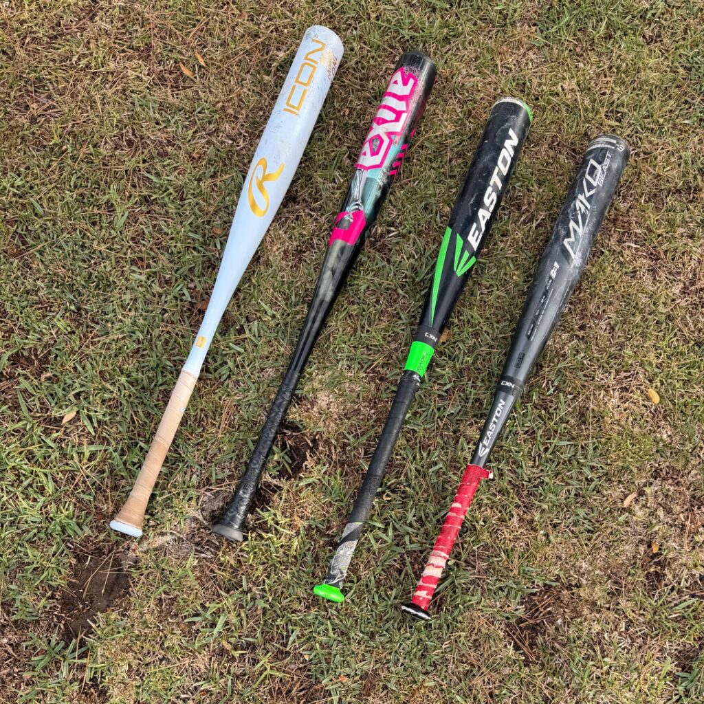 Four high school BBCOR baseball bats lying on the grass, showing the mandatory and often expensive equipment upgrade for 14U and older players.