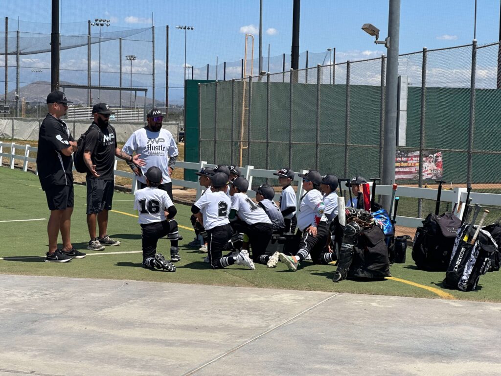 Three coaches standing and talking to a kneeling MVP Hustle 8U travel baseball team outside the dugout next to their equipment bags