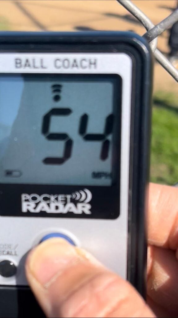 Pocket Radar displaying a 54 mph pitching velocity through a fence, typical for a 12U AA travel baseball team.
