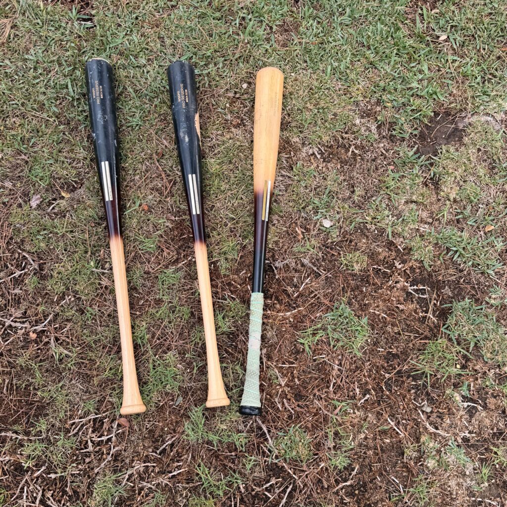 Three Warstic wood baseball bats lying on the grass, a recurring equipment expense for high school travel ball tournaments.