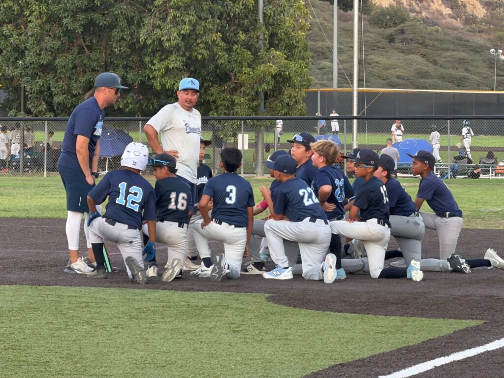 Two coaches talking to a kneeling ZT Prospects youth travel baseball team during a postgame huddle on a turf field.